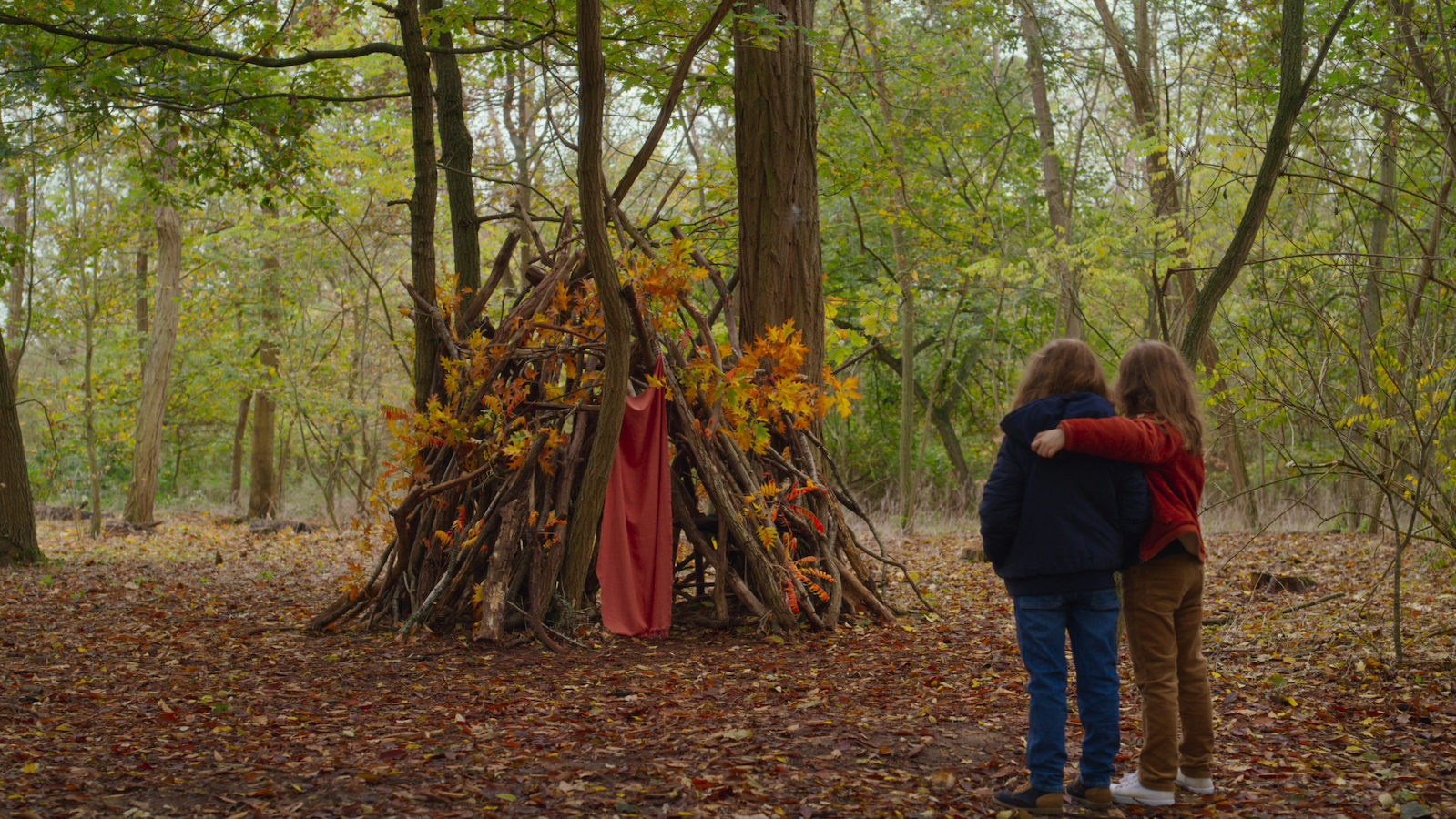 Two little girls seen from behind in the wood looking at a fort made of sticks