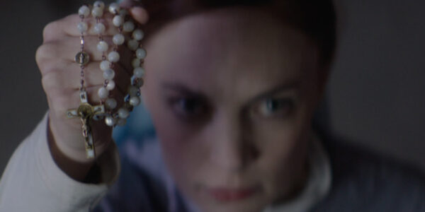 A woman holds up a pearly necklace towards the camera in close-up, her face blurred and out of focus
