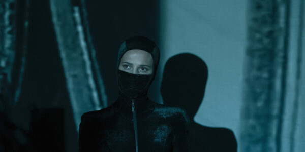 A woman in a zip-up black leather suit and the bottom of her face obscured by a mask looks past the camera; her shadow on the wall behind her