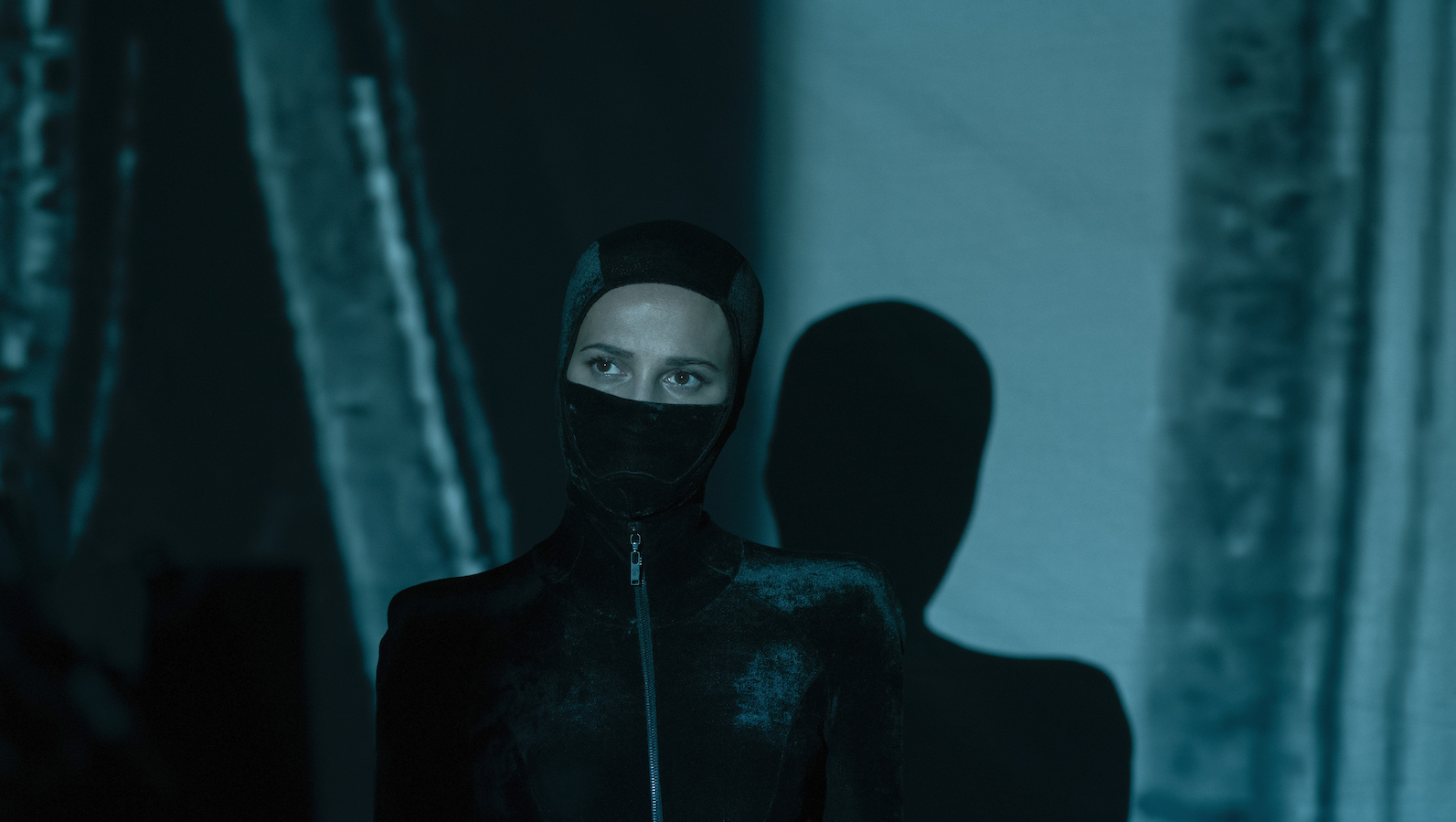 A woman in a zip-up black leather suit and the bottom of her face obscured by a mask looks past the camera; her shadow on the wall behind her