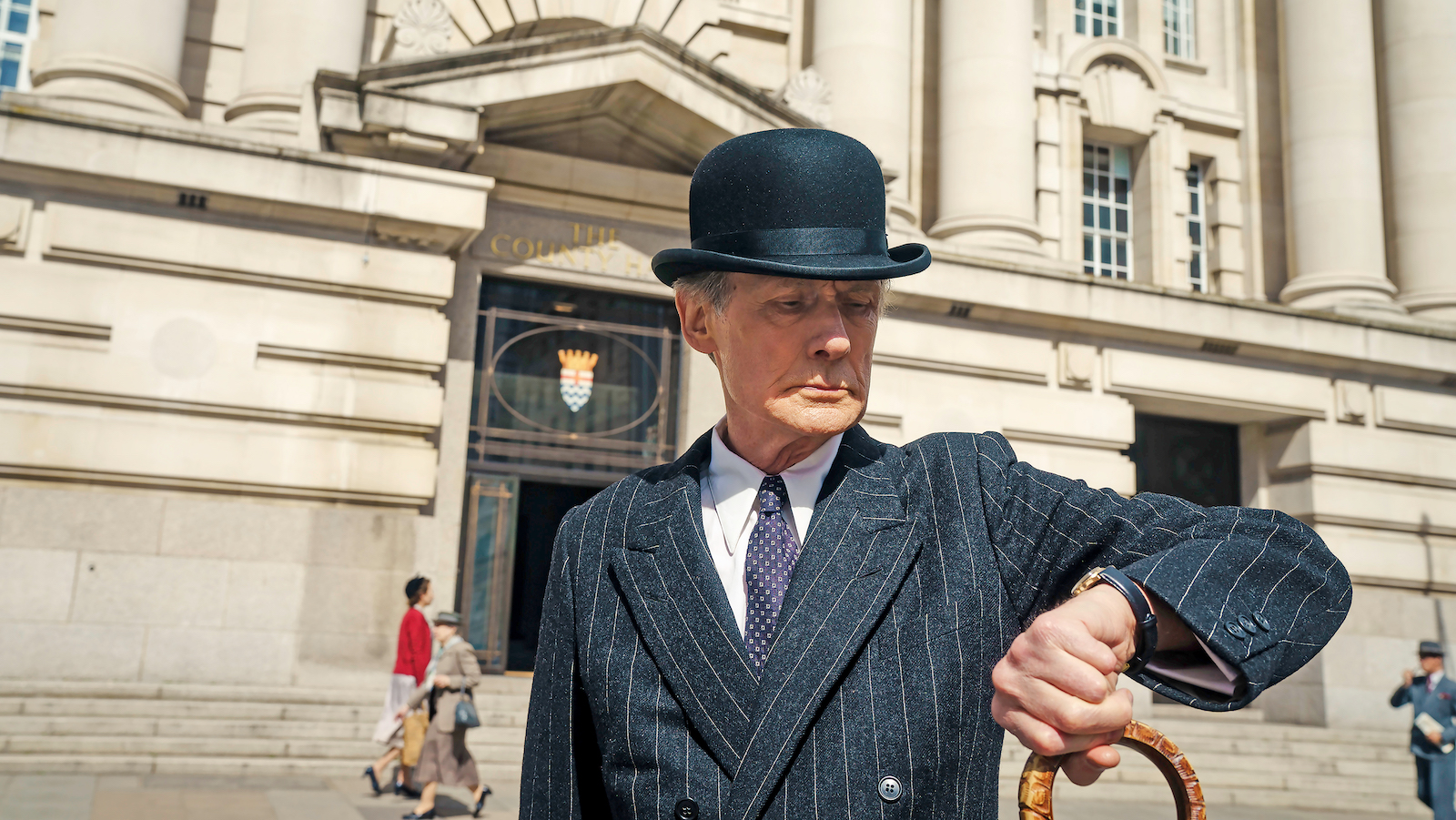 A man in a bowler hat stands outside a building looking at his watch
