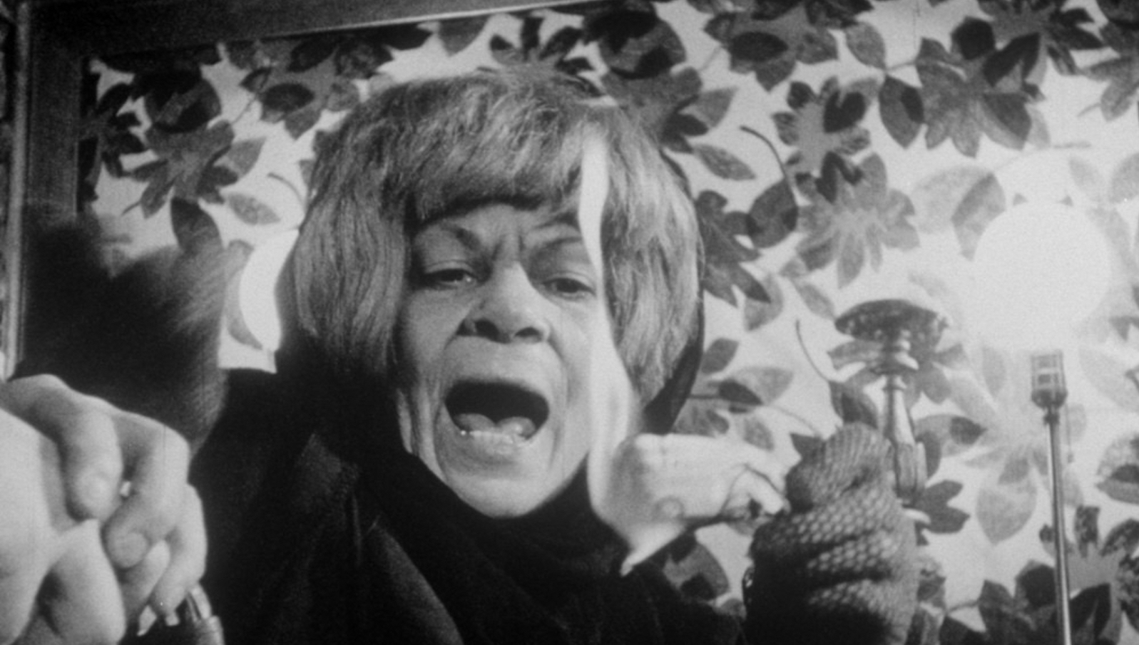 An older woman with gray hair holds a burning piece of paper, her moth agape