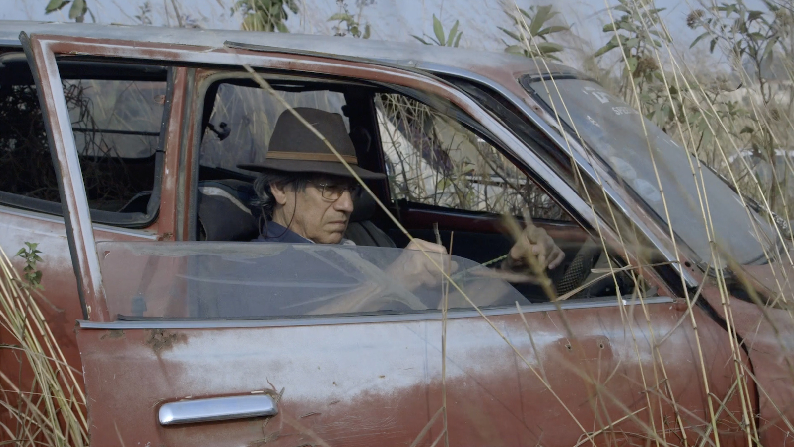 A man sits in a stopped car's passenger seat, the door open, surrounded by tall grass and weeds