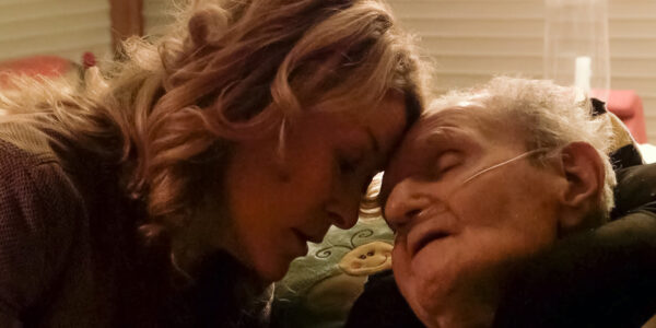 A woman rests her forehead against an elderly man's forehead as he lies in bed, his mouth open and eyes closed.