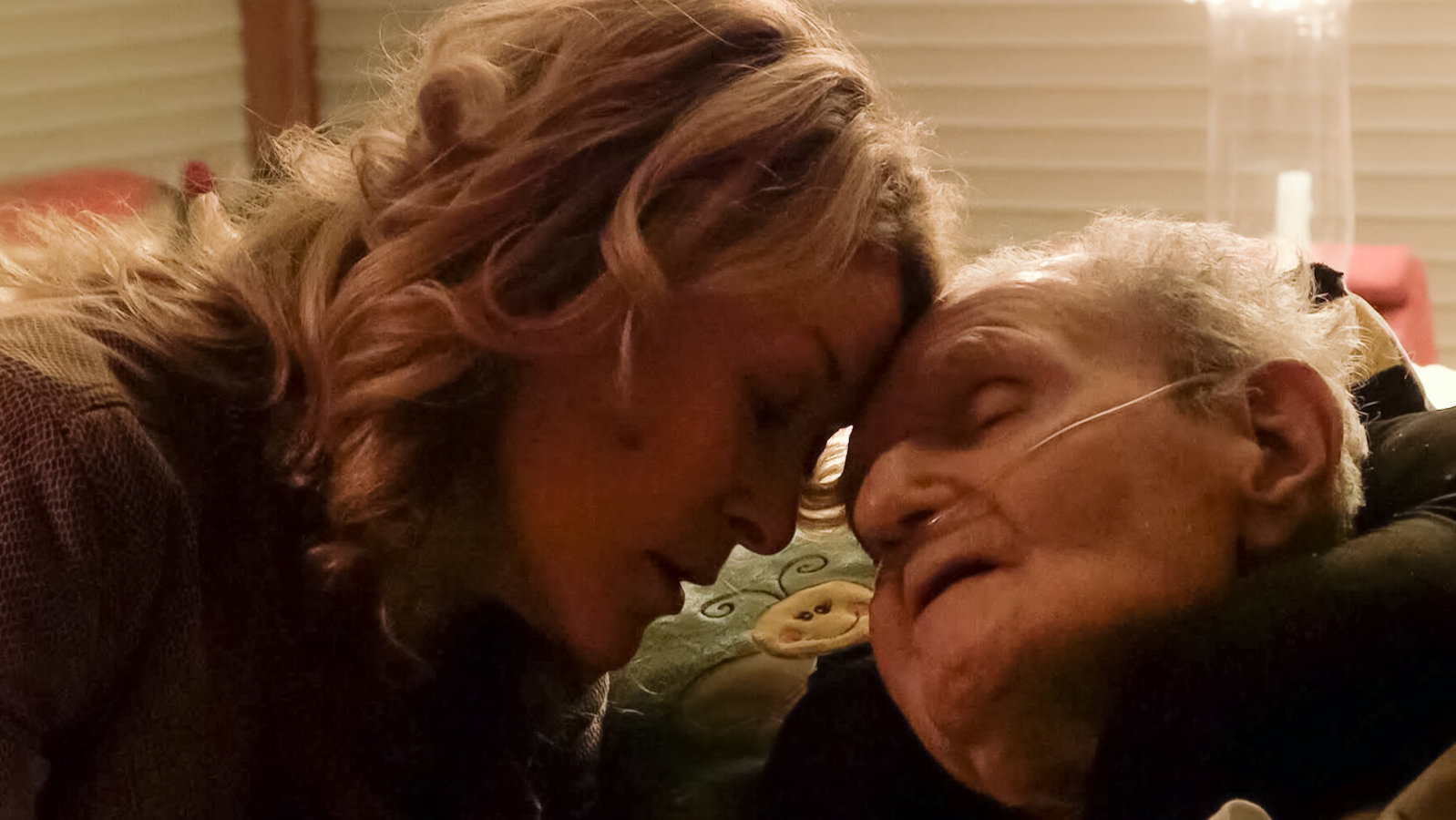 A woman rests her forehead against an elderly man's forehead as he lies in bed, his mouth open and eyes closed.