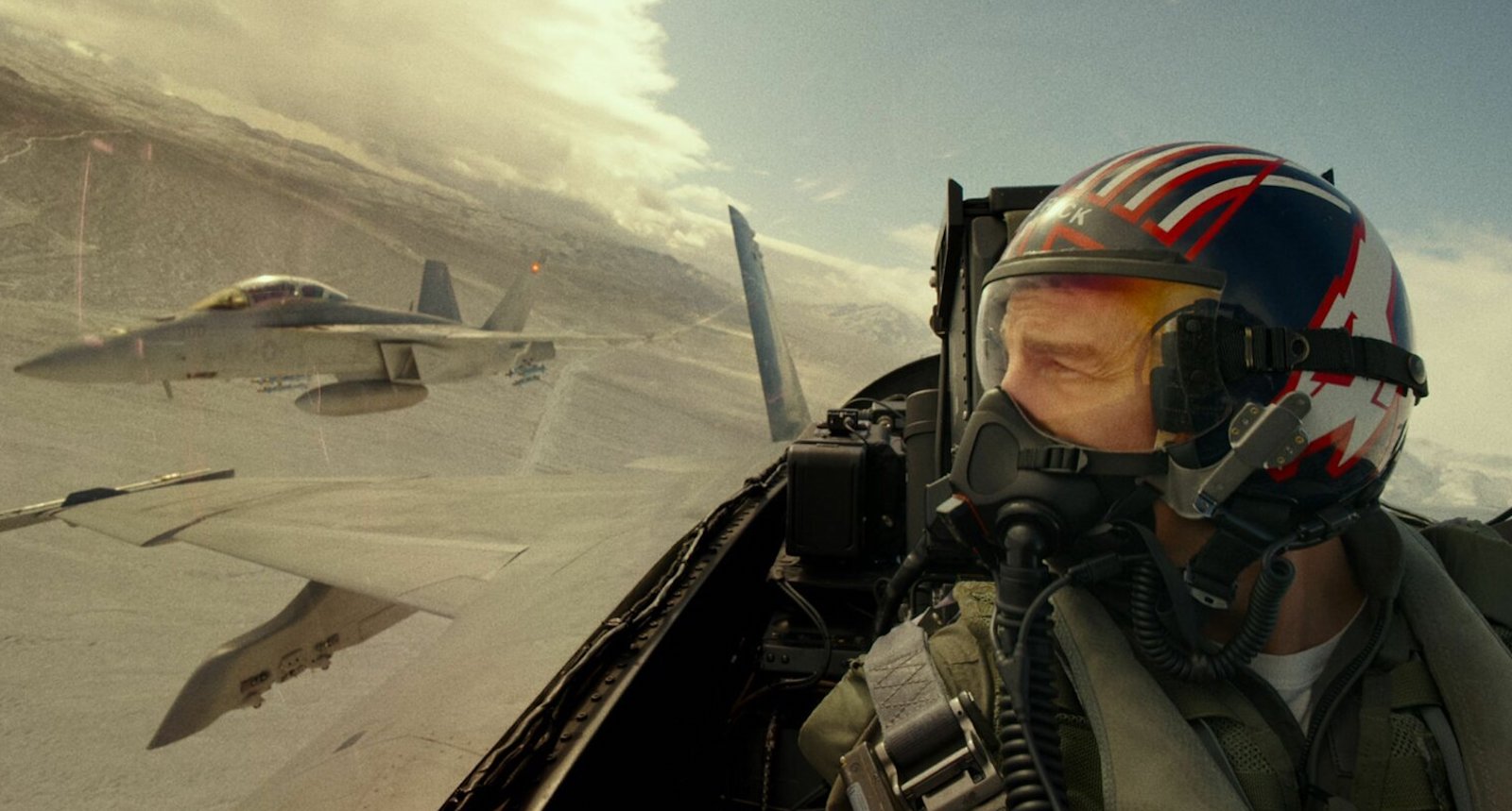 A navy pilot in helmet and mask flies in his cockpit while enemy planes fly close-by in the sky to his left