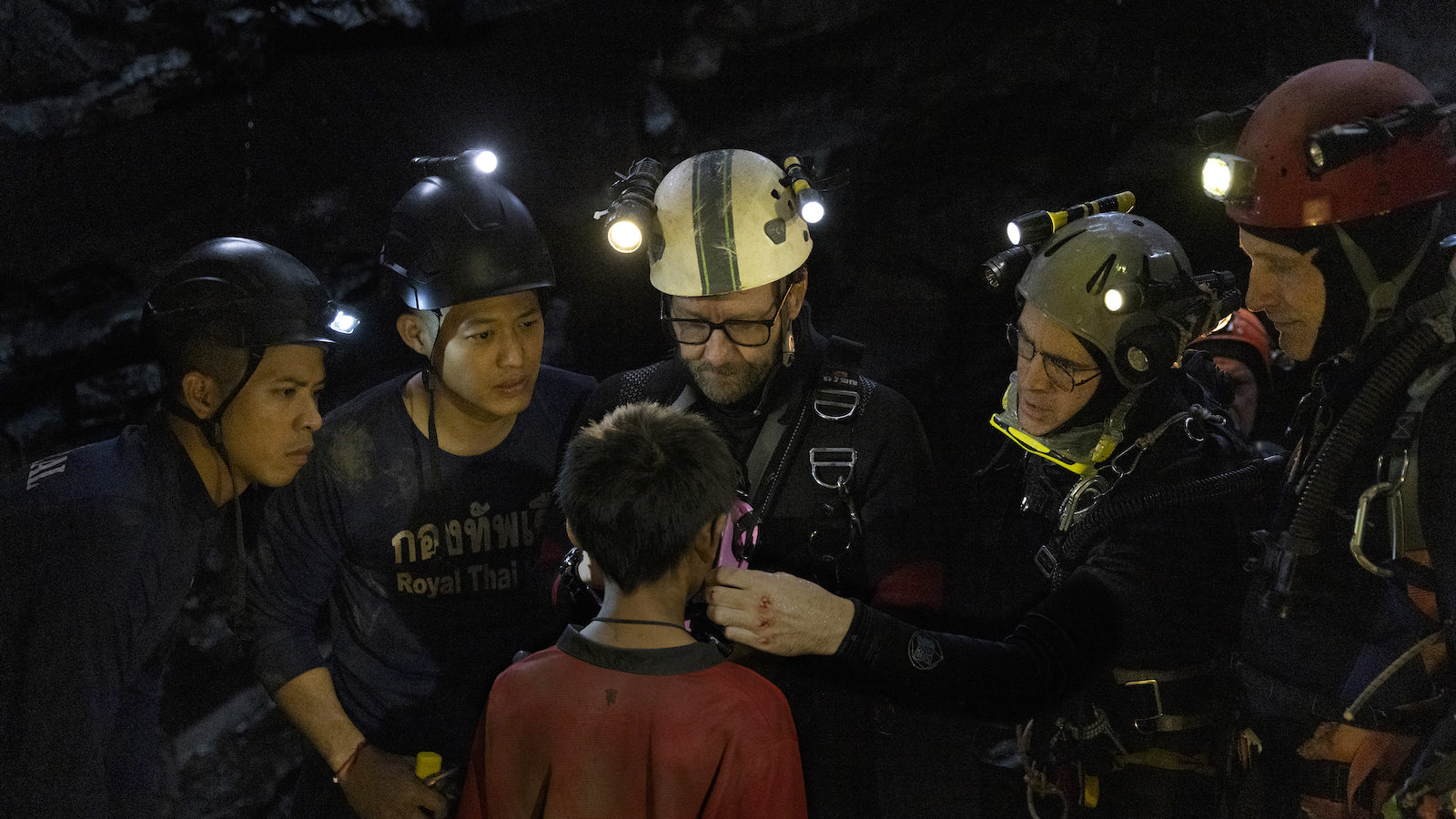 In a cave, a group of divers in helmets stand around a boy, seen from the back, in a red shirt.