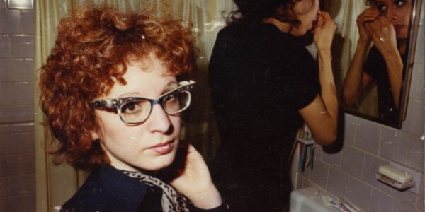 A woman in glasses and frizzy red hair looks in the camera away from a mirror