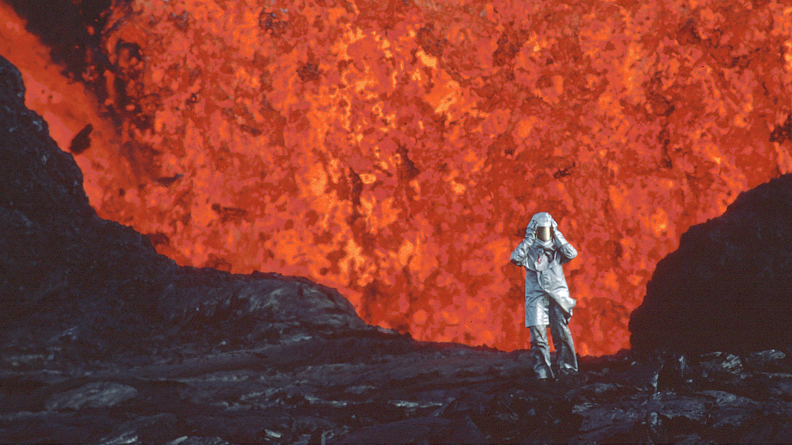 A woman wearing an aluminized suit standing near a red lava burst.