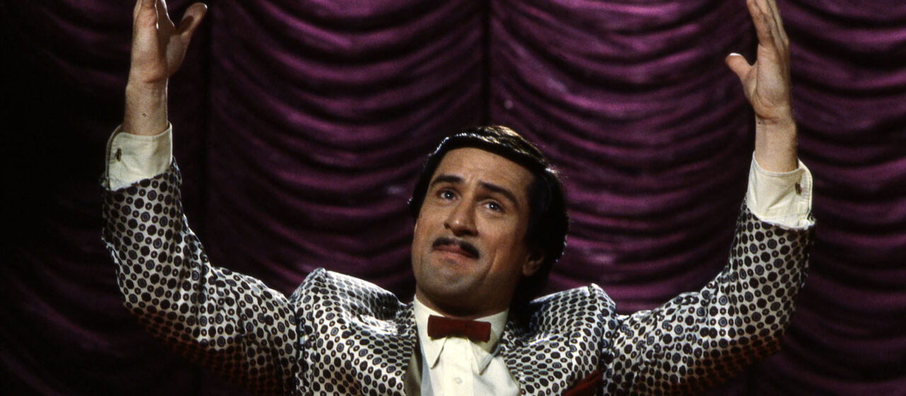 A mustachioed man with his hands up standing in front of a velvet curtain on stage