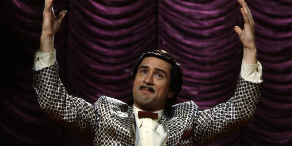 A mustachioed man with his hands up standing in front of a velvet curtain on stage