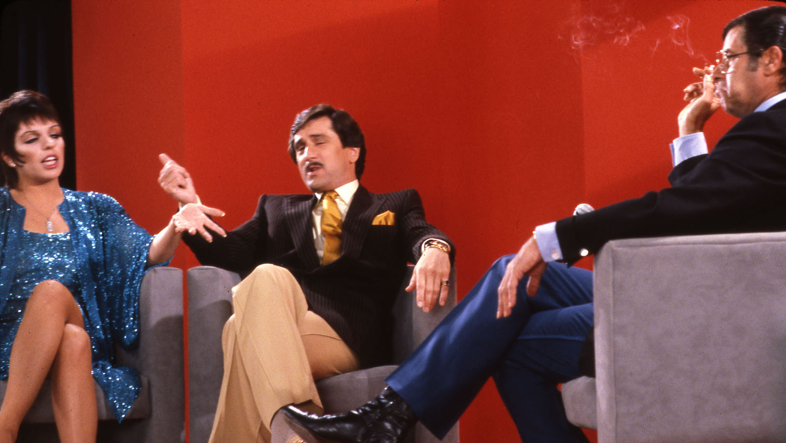 Three people sit on a talk show set with a red background