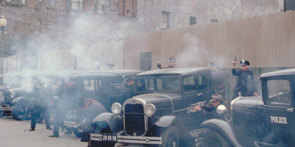 Cops firing guns have a shootout with off-screen forces in a parking lot with cars from the 1920s