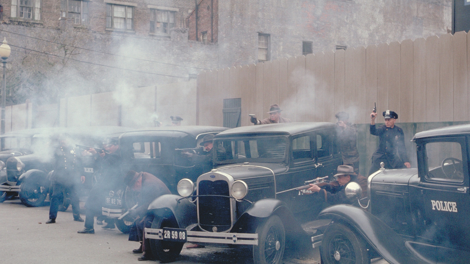 Cops firing guns have a shootout with off-screen forces in a parking lot with cars from the 1920s