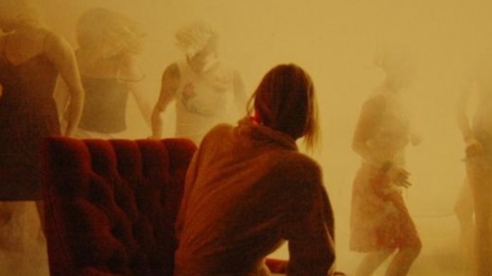 A woman seen from the back sits in a smokey yellow room and watches other women dance in the fog