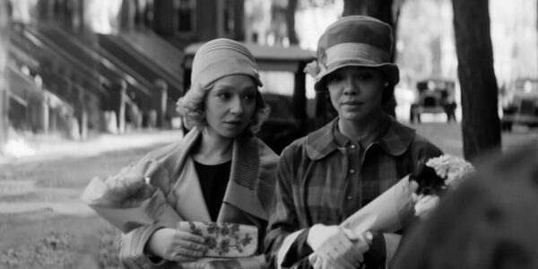 A black and white image of two women talking while walking down a New York street in the 1920s, wearing hats and gloves