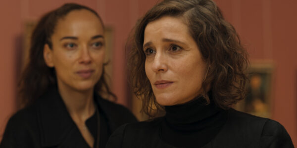 Two women stand, one in foreground looks off camera to the right, the other looking at her