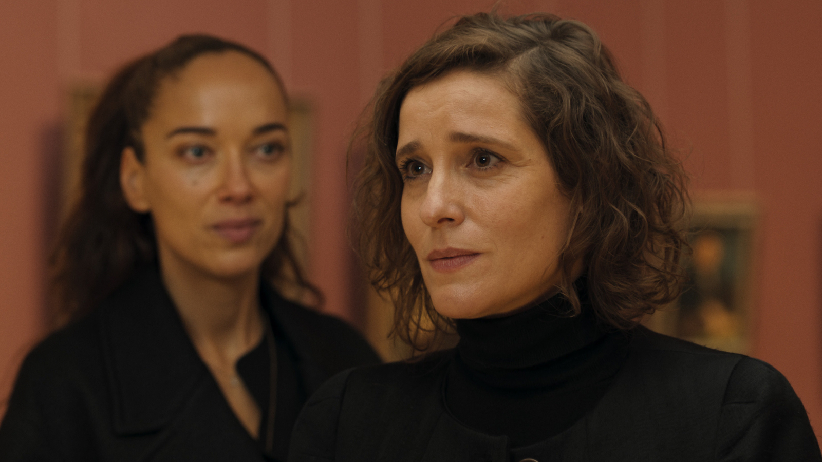Two women stand, one in foreground looks off camera to the right, the other looking at her