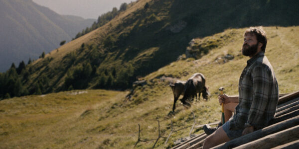A man sits on a rooftop and looks out at a mountainous landscape, with horses grazing behind him