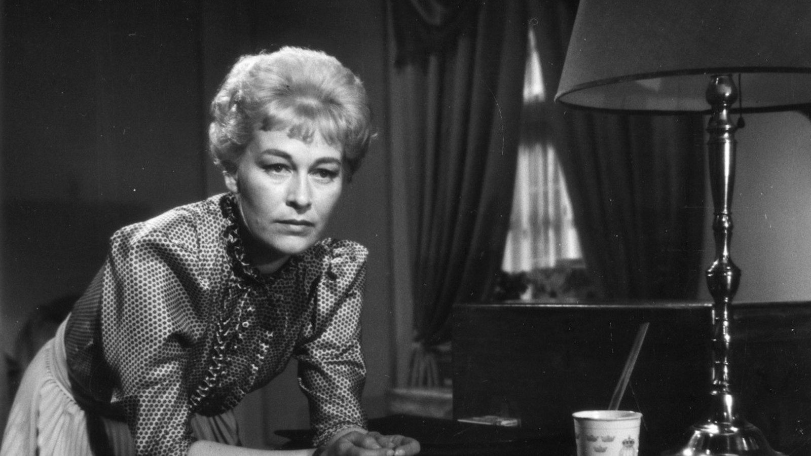 A woman with short blonde hair rests on her elbows expectantly, next to a lamp