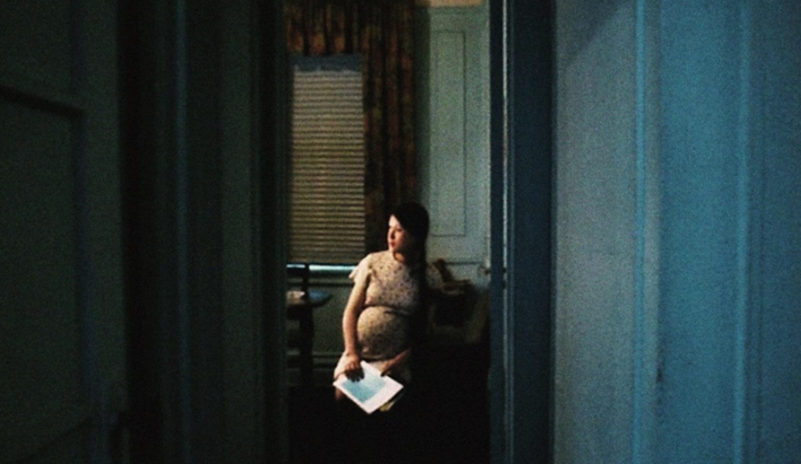 a pregnant woman sits alone, holding a piece of paper in a vacant room, shot from a distance