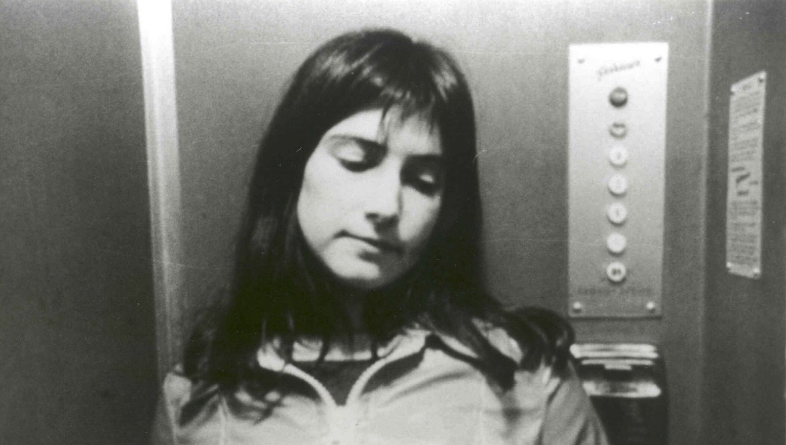 A woman with long dark hair stands in an elevator looking down