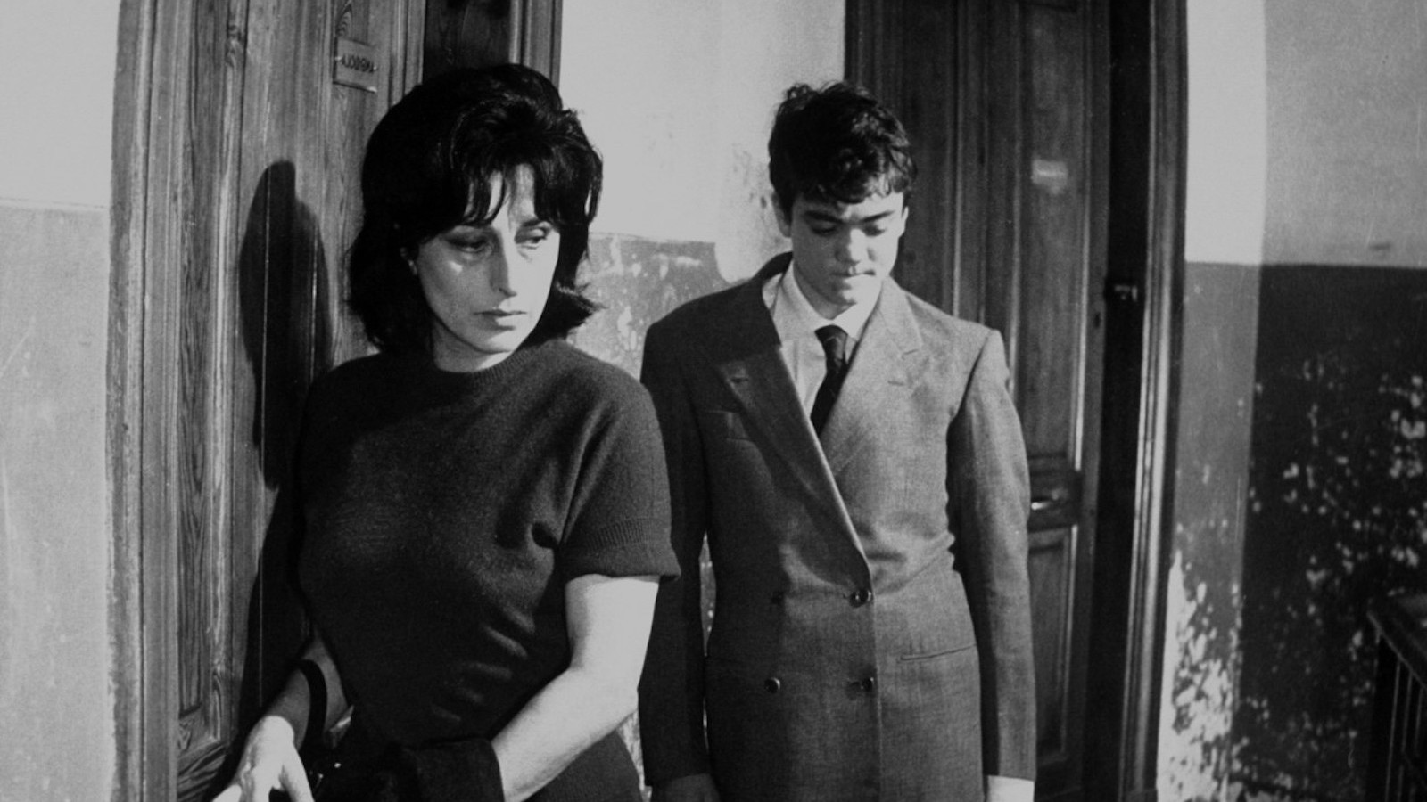 A middle aged woman with dark hair and her son with dark hair stand in a room, he slightly behind her