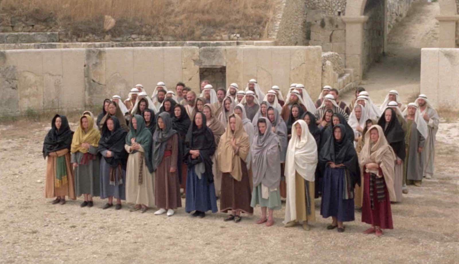 A group of people in Biblical clothing and shawls stand in a sunny desert area, lined up in single file in four rows