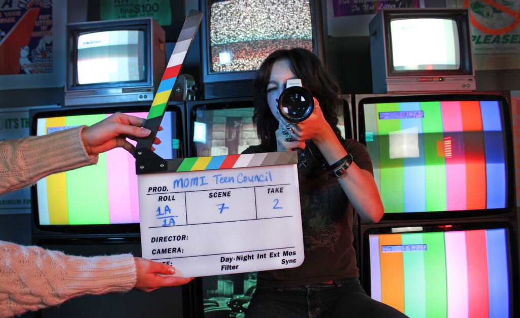 Teen Council A young woman holds a camera aimed at the screen and stands behind a slate and clapper