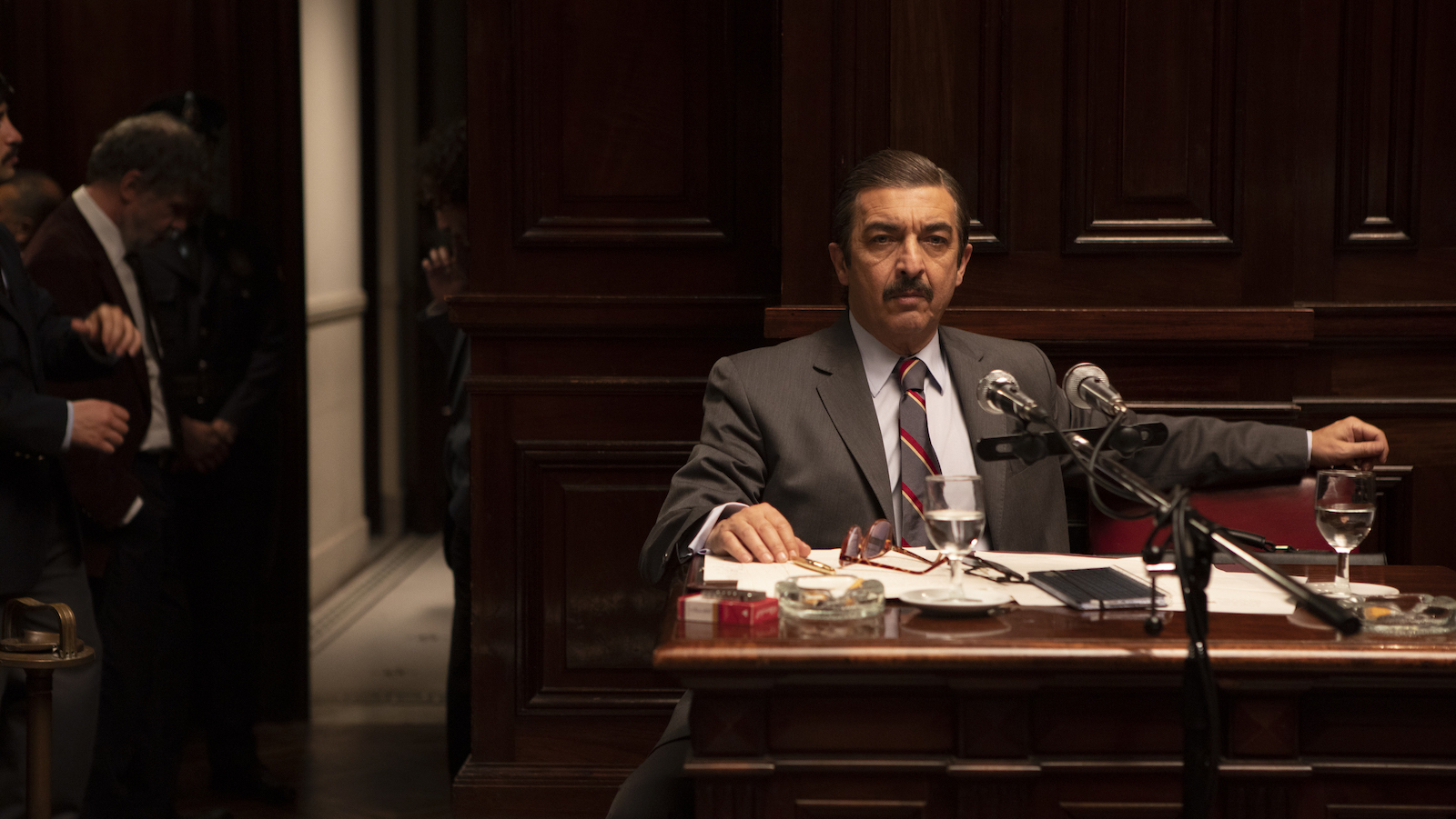 A mustachioed man sits in a dark room with wood paneling in front of a microphone, lit from above
