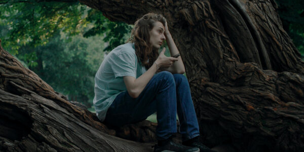 A young person with long hair sits on a tree in a forest holding a phone and looking into the distance