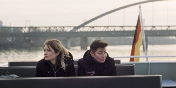 Two women sit on a ferry, huddled in the cold, a bridge in the background