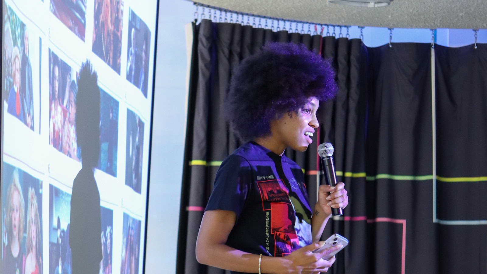 A woman talks into a microphone standing in front of a screen as she reads trivia questions
