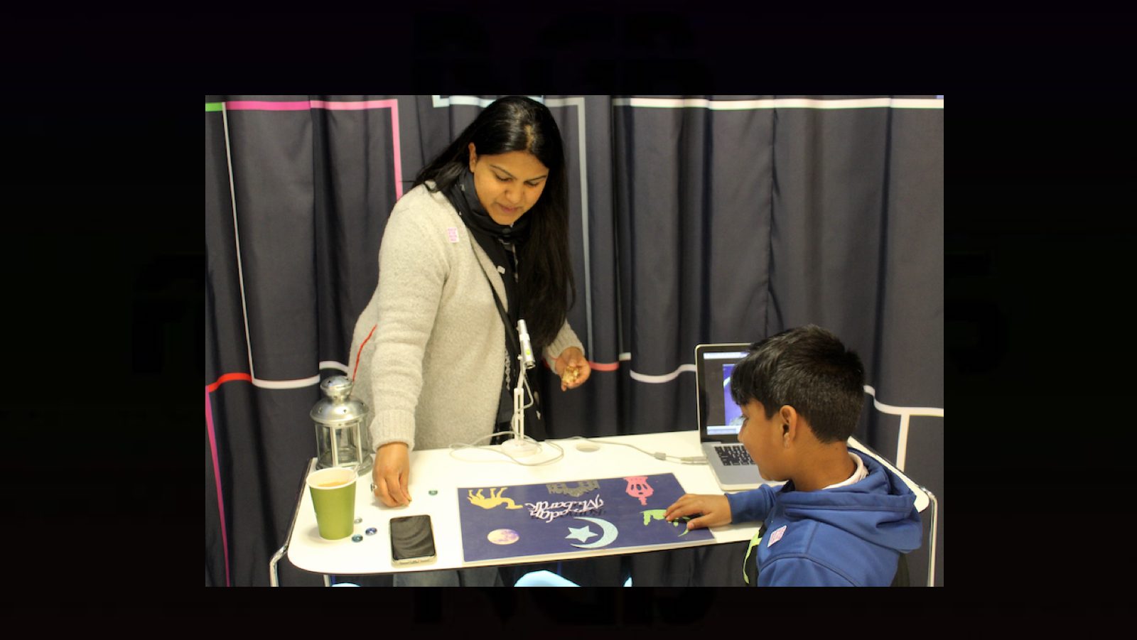 A teacher stands in front of a work table, instructing a child with construction paper and cut outs