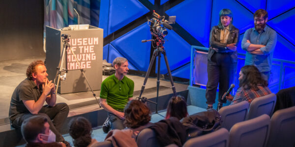 Two men sit on the edge of a stage with filmmakers and a camera to their left and a group of people in the front row, their backs to the camera.