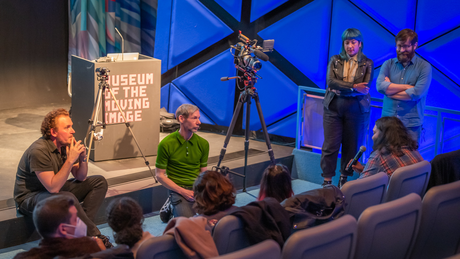 Two men sit on the edge of a stage with filmmakers and a camera to their left and a group of people in the front row, their backs to the camera.