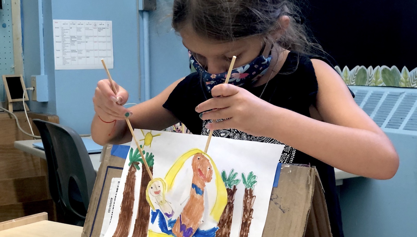 A masked child constructs paper puppets with wooden rods.