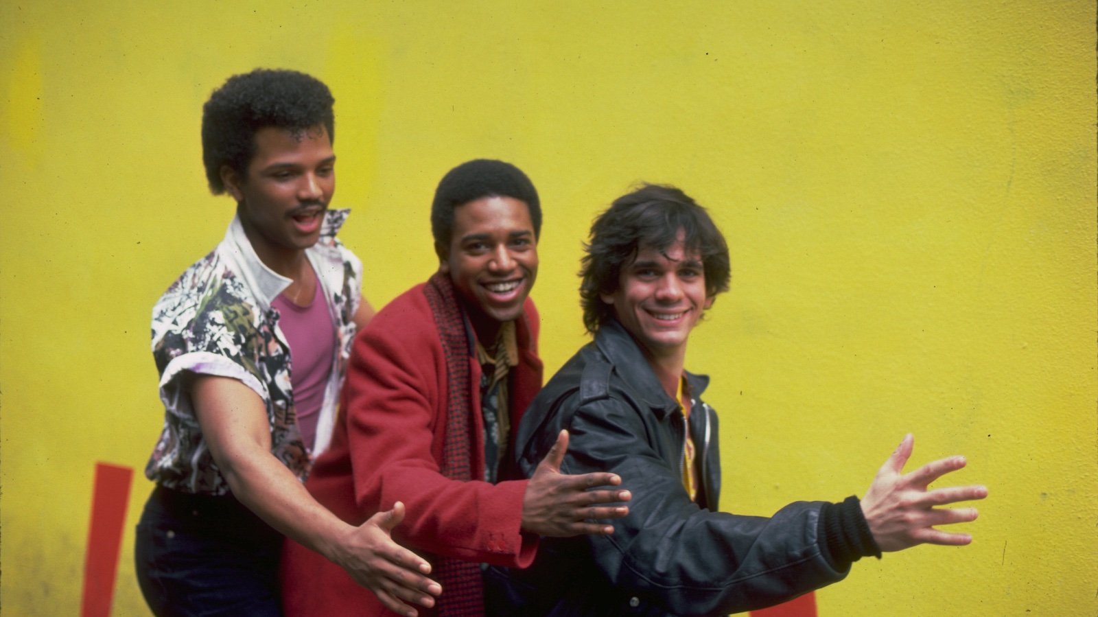 Three men smile and pose in front of a yellow background