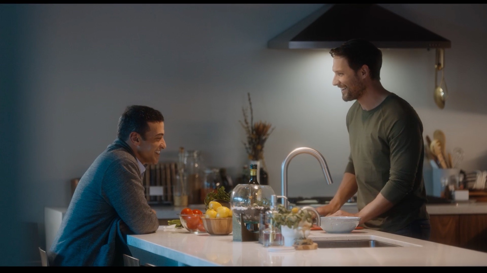 Two men in a kitchen, one sitting in front of an island, the other standing over the island, his head under a ceiling lamp