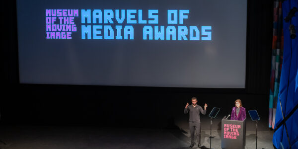 a white man in a sparkly purple suit stands at a podium on a large stage with a sign-language interpreter next to him while on the screen there are words that read Marvels of Media Awards