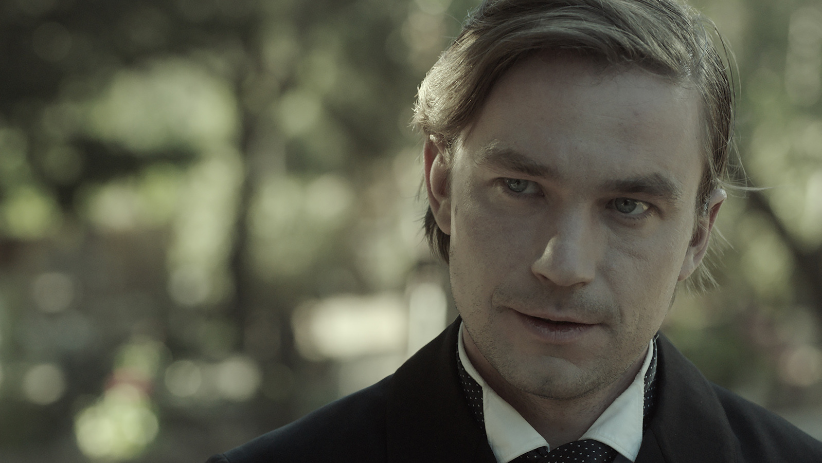 A blond man in medium close-up smirks and looks past the camera
