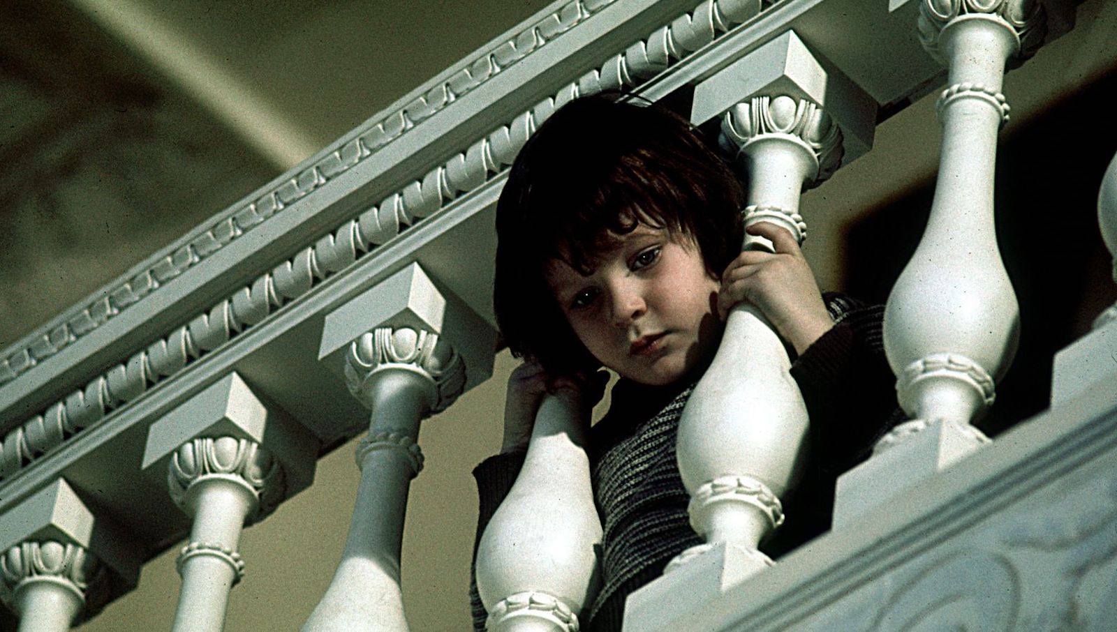 A little mop haired boy pokes his head through staircase railings and looks down