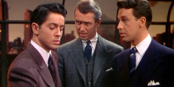 Three men stand in a room by a large window during sunset, all three wear suits, while the older man in the middle looks intently at the man on the left, who looks away painfully
