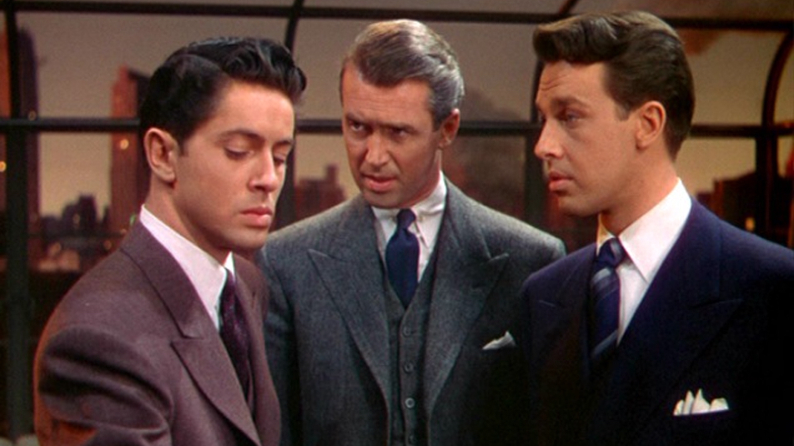 Rope Three men stand in a room by a large window during sunset, all three wear suits, while the older man in the middle looks intently at the man on the left, who looks away painfully