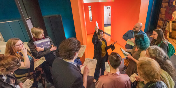 A tour guide holds up an object among a crowd of visitors in the Museum's galleries