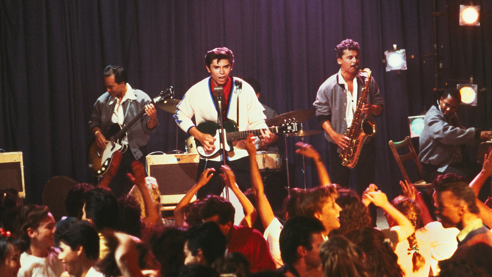 A man in a white sweater sings on stage with a guitar.