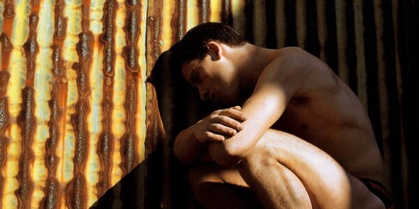 A naked man crouches on the ground near a rusted metal fence