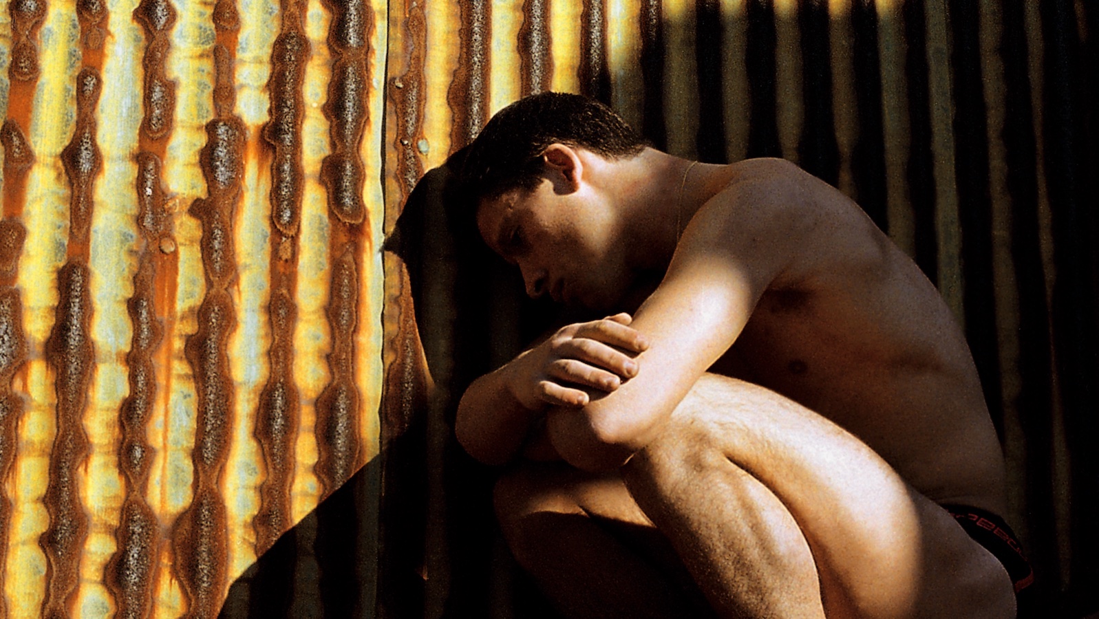 A naked man crouches on the ground near a rusted metal fence
