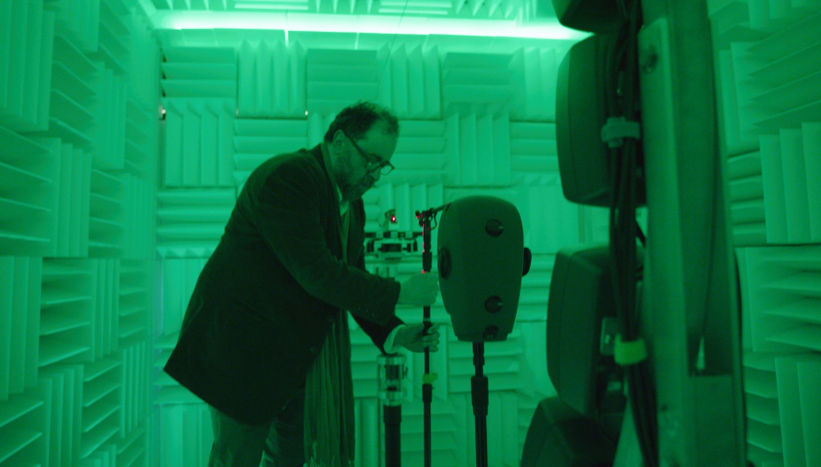 A man in glasses holds a microphone stand in a sound-proofed room all drenched in green light