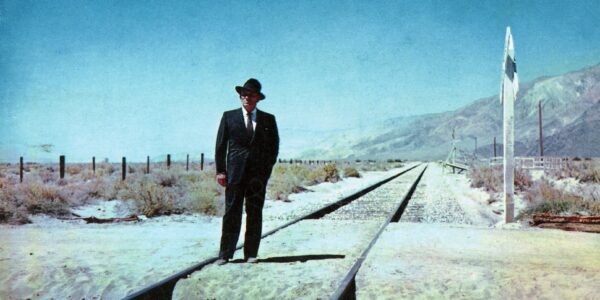 A one-armed man in a black suit and fedora stands on a railroad track in the middle of nowhere against a blue sky